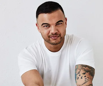 Man with short dark hair and arm tattoos wearing a white t-shirt, smiling against a light background.