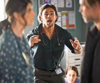 Young man with stethoscope urgently gestures in a hospital setting, others partially visible.