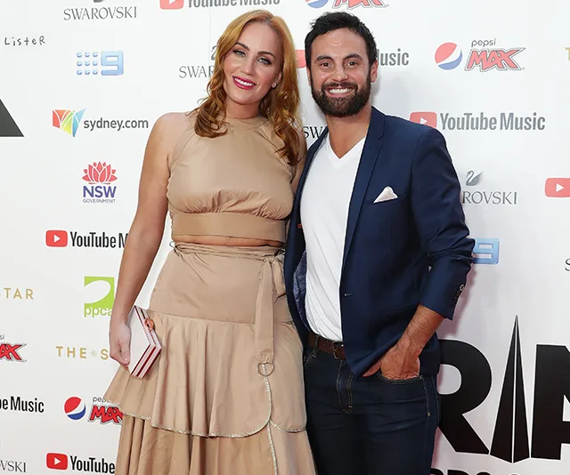 A couple posing on a red carpet; the woman is wearing a beige dress and the man a navy blazer with jeans.