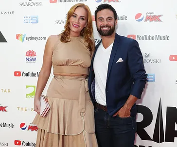 A couple posing on a red carpet; the woman is wearing a beige dress and the man a navy blazer with jeans.