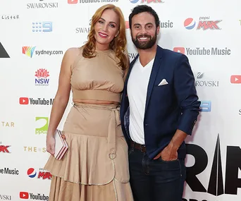 A couple posing on a red carpet; the woman is wearing a beige dress and the man a navy blazer with jeans.