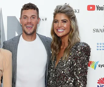 A smiling man in a gray blazer stands next to a woman in a sequined dress at a promotional event backdrop.
