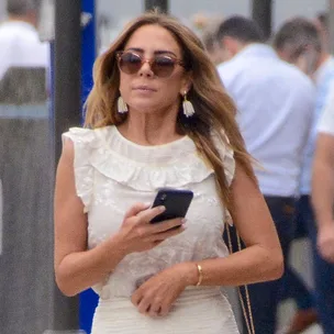 Woman in white dress and sunglasses walking outdoors, holding a phone.