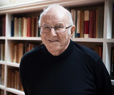 Elderly man with glasses and a black sweater stands smiling in front of bookshelves.