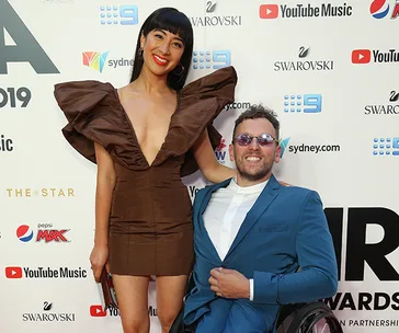 Two individuals pose at a red carpet event, one in a wheelchair, both smiling.