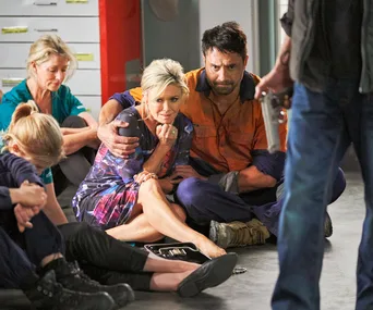 "Scene from Home and Away, characters sitting on the floor, one holds another as a person with a gun stands in front."