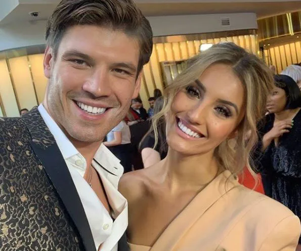 Couple smiling at an event, with the man in patterned blazer and the woman in a cream outfit, surrounded by people.