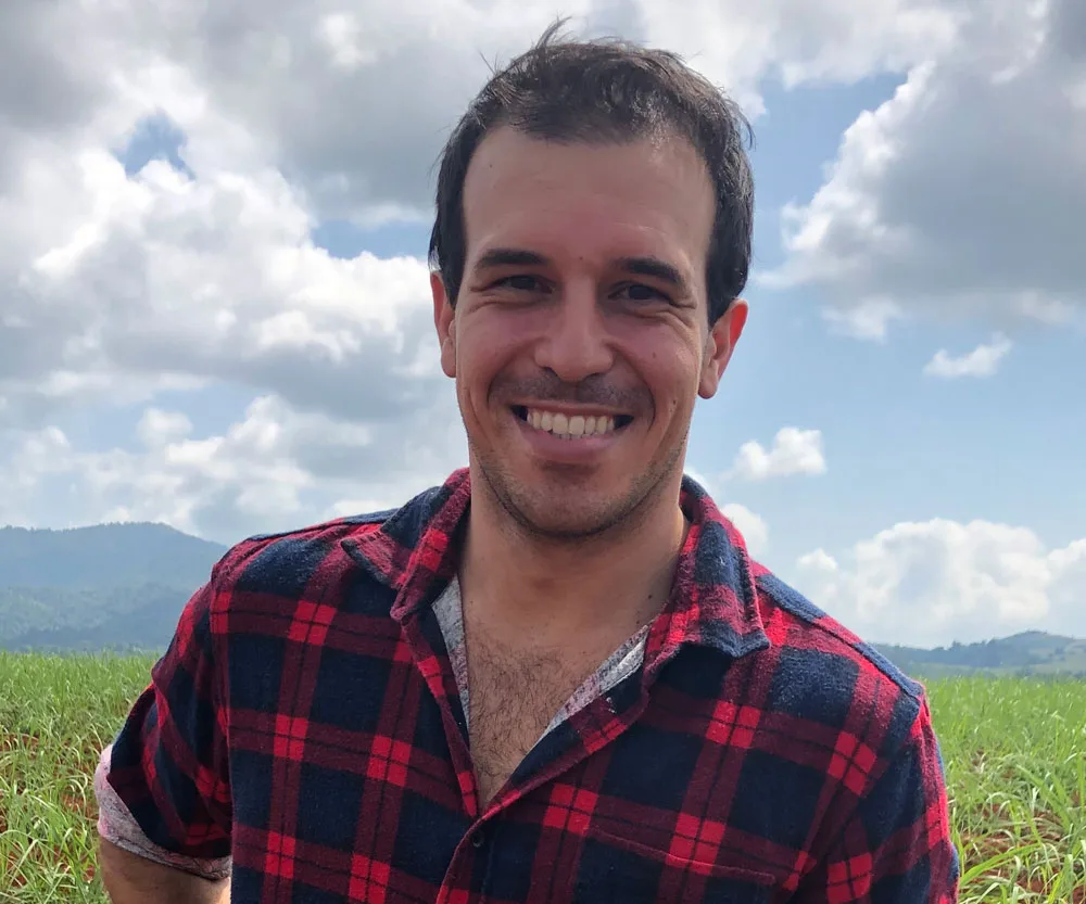 Man in red plaid shirt smiles in a scenic rural field, under a partly cloudy sky.