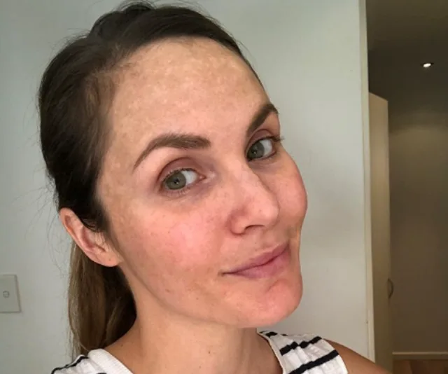A woman with light skin and dark hair tied back, wearing a striped tank top, looks at the camera in a well-lit room.