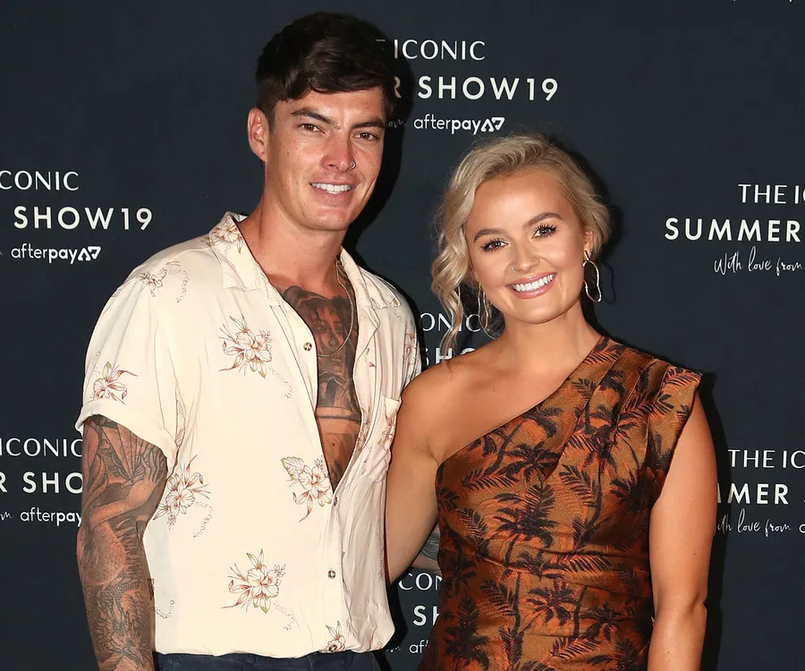 Two individuals smiling at a fashion event, posing in front of a branded backdrop.