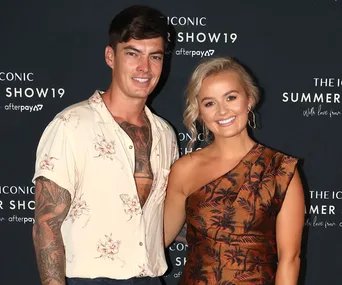 Two individuals smiling at a fashion event, posing in front of a branded backdrop.