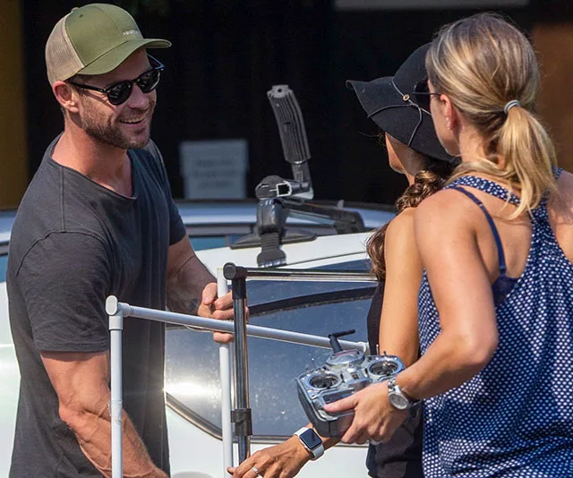 Person in sunglasses and cap interacts with two women by a car, holding equipment outdoors on a sunny day.