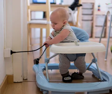 Baby in a walker pulling an electrical cord from a socket, creating a potential safety hazard in a home setting.