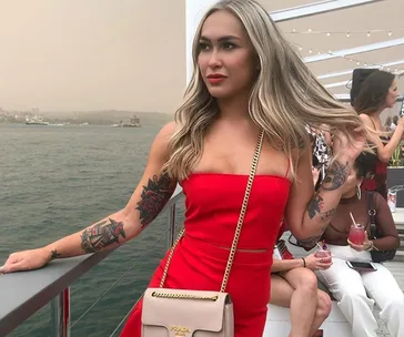 Woman in red dress with tattoos, standing on a balcony by the water, holding a beige bag.