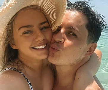 A couple posing happily on a sunny beach, with the woman in a sun hat and the man giving a playful kiss.