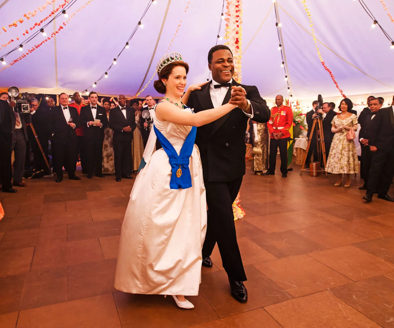 Elegant woman in a white gown and tiara dances with a man in a tuxedo under a tent with string lights, surrounded by onlookers.