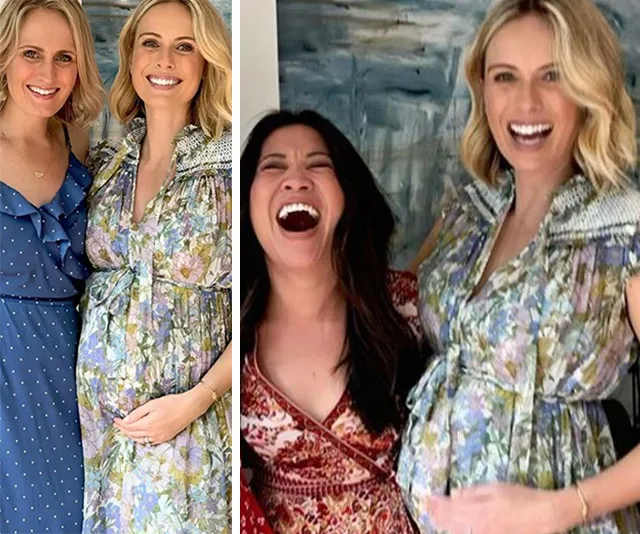 Two women smiling at a baby shower, one in a floral dress touching her baby bump, the other in blue standing beside her.