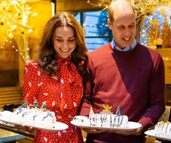 Two people smiling while holding festive Christmas cakes indoors with holiday decorations around.