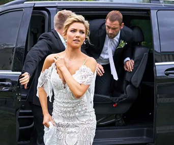 A woman in a white wedding dress hurriedly exits a black vehicle, followed by a man in a suit.