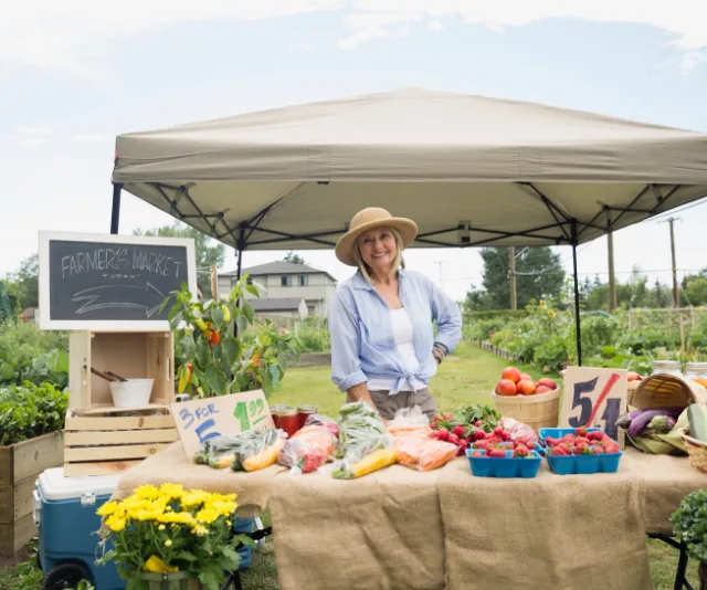 Farmer's market vendor smiling under a canopy with fresh produce on display, including vegetables and flowers.