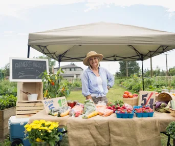 Farmer's market vendor smiling under a canopy with fresh produce on display, including vegetables and flowers.