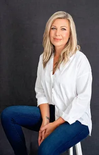 A woman with long blonde hair, wearing a white shirt and blue jeans, seated against a dark backdrop.