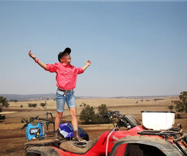 A child in a pink shirt and cap joyfully stands on an ATV in an open, dry landscape.
