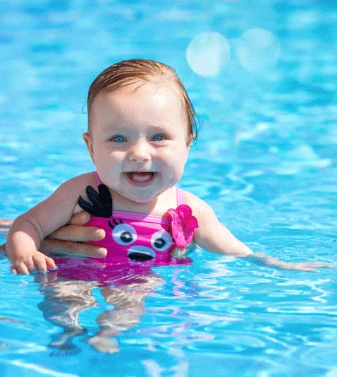 A baby with a pink swimsuit is held in a pool, smiling happily.