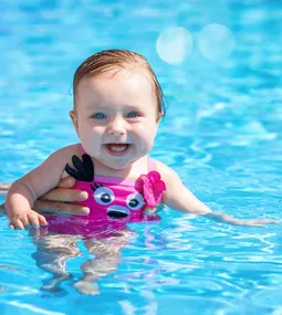 A baby with a pink swimsuit is held in a pool, smiling happily.