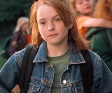 Young girl with red hair wearing a denim jacket and backpack, standing outdoors with other kids in the background.