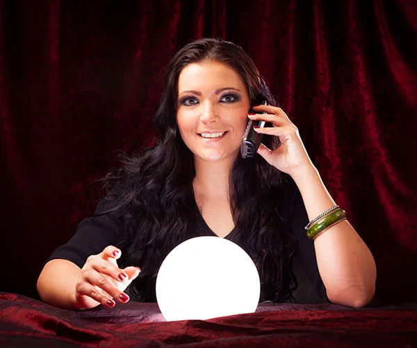 A woman with long dark hair holds a phone and sits behind a glowing crystal ball, with a red velvet curtain in the background.
