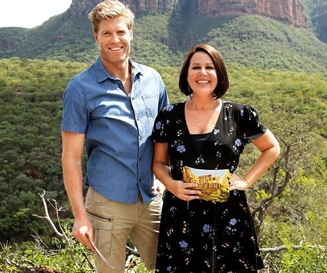 Two people smiling outdoors against a mountain landscape, one holds a "Celebrity Get Me Out of Here" envelope.