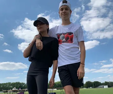 Mother and son pose outdoors on a sunny day; she wears black, he wears a white T-shirt.