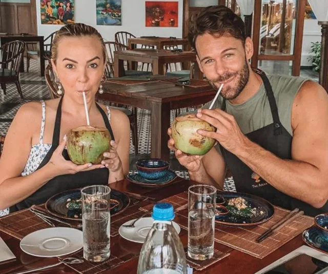 Two people sitting at a table in a restaurant, drinking from coconuts with straws and smiling at the camera.