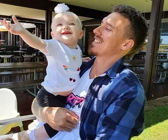 A smiling man holds a joyful baby wearing a white shirt and headband, reaching out in an outdoor setting.