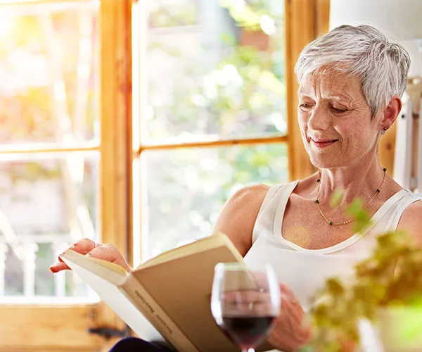 Elderly woman reading a book by a sunny window with a glass of red wine beside her.