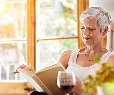 Elderly woman reading a book by a sunny window with a glass of red wine beside her.