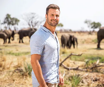 Man in a light shirt stands in a savanna with elephants in the background.