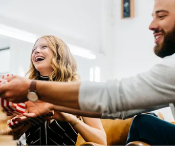 Two people exchanging a gift, with one person laughing joyfully, in a bright indoor setting.