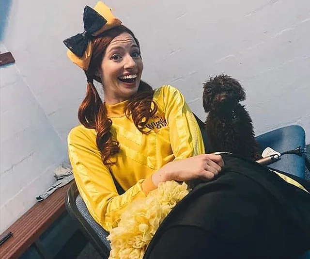 Woman in a yellow costume with a large bow, smiling and sitting on a chair, next to a small brown poodle.