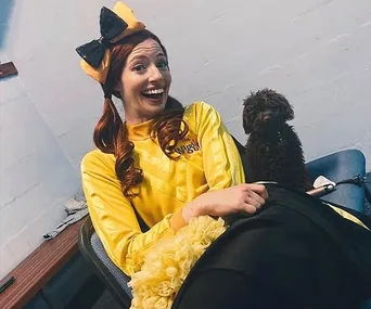 Woman in a yellow costume with a large bow, smiling and sitting on a chair, next to a small brown poodle.