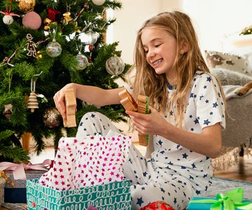 Young girl in star-patterned pajamas joyfully opening Christmas gifts by a decorated tree.