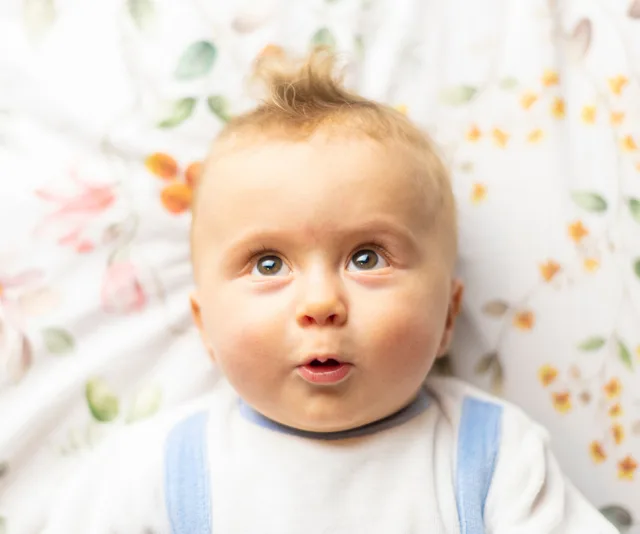 Baby lying on floral bedspread, looking upward with a surprised expression and wearing a white outfit with blue straps.
