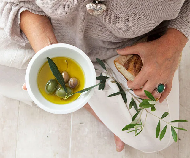 Person holding a bowl of olive oil with olives and a piece of bread.