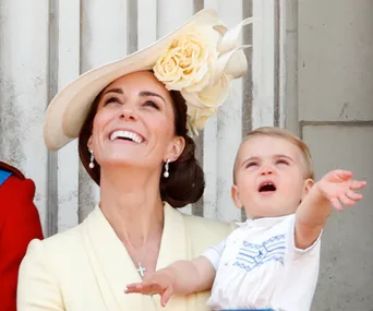 A woman in a cream outfit and hat with a large flower smiles, holding a young child pointing upwards against a stone backdrop.