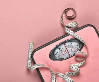 A pink bathroom scale with a coiled measuring tape on a pink background.
