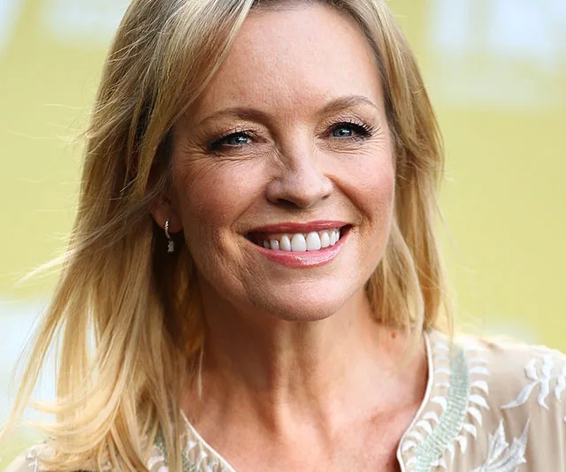 A woman with blonde hair smiling, wearing a light-colored top with a detailed pattern.