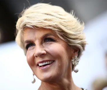 A woman with short blonde hair, smiling, wearing pearl earrings at an event.