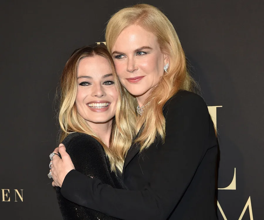 Two women with blonde hair pose together, one embracing the other, smiling in front of a dark background.