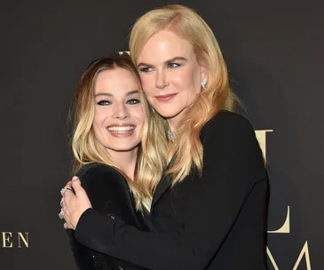 Two women with blonde hair pose together, one embracing the other, smiling in front of a dark background.